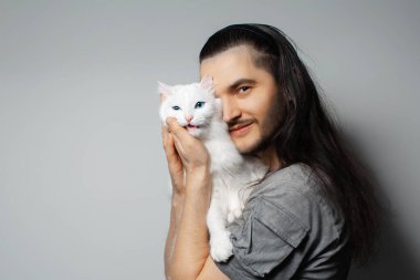 Gri arka planda beyaz kedisi olan genç bir adamın stüdyo portresi. Mavi gözlü evcil hayvan.
