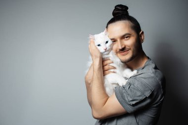 Gri arka planda beyaz kedisi olan genç bir adamın stüdyo portresi. Mavi gözlü evcil hayvan.