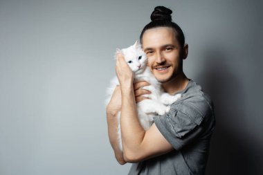 Gri arka planda beyaz kedisi olan genç bir adamın stüdyo portresi. Mavi gözlü evcil hayvan.