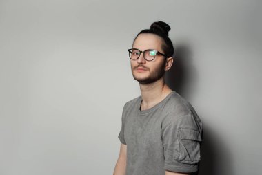 Saçları topuzlu, gözlüklü ve gri arka planda gömlek giyen genç bir adamın stüdyo portresi..