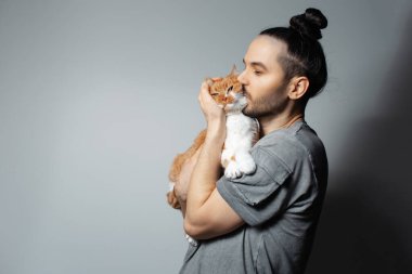 Stüdyo portresi. Kucağında kırmızı kedi olan genç adam. Gri arka planda evcil hayvanı öperken..
