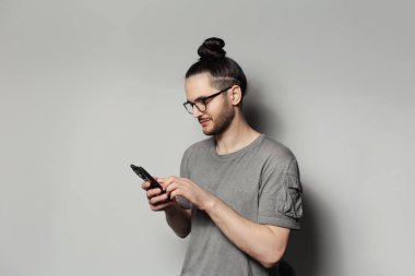 Stüdyo portresi, saçı topuzlu bir gencin gri arka planda akıllı telefon ekranına yazı yazması..