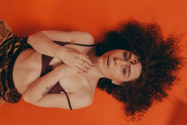 Stüdyodaki kızıl saçlı kıvırcık kız turuncu arka planda şık kıyafetler giyiyor.