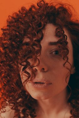 Stüdyodaki kızıl saçlı kıvırcık kız turuncu arka planda şık kıyafetler giyiyor.