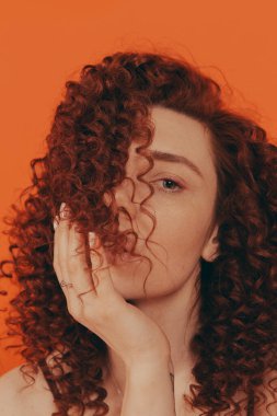 Stüdyodaki kızıl saçlı kıvırcık kız turuncu arka planda şık kıyafetler giyiyor.