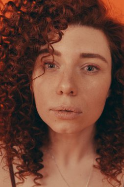 Stüdyodaki kızıl saçlı kıvırcık kız turuncu arka planda şık kıyafetler giyiyor.