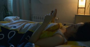 Uykusuzluk çeken, interneti kolaçan eden, uyumadan önce akıllı telefondan video izleyen kadın. Yatağa uzanmış kedi. Uyku Bozukluğu. 4K numaralı telefondan mesajlar ve e- postalar kontrol ediliyor