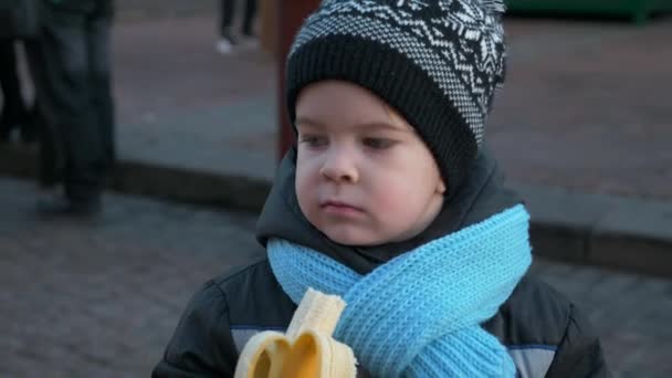 饥饿的孤独儿童在圣诞市场独自站着吃香蕉 夜城悲伤的情感严肃的男孩的脸 2X慢动作60 Fps — 图库视频影像