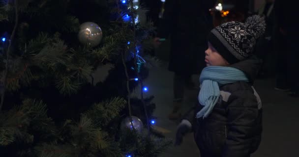 クリスマスツリー シンキング ドリーミングの近くに深刻な子供が立っている 見てる 夜市駅を歩いている人たち — ストック動画
