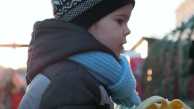 Aç Yalnız Çocuk, Noel Pazarı 'nda Tek Başına Muz Yiyor. Kış tatilleri. Akşam Şehri. Üzgün Duygular Ciddi Erkek Yüzü. 2x Yavaş çekim 60 FPS 4K