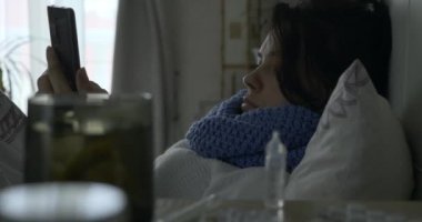 Hasta Kadın Smartphone Kullanıyor İnternet Tarayıcı Dokunmatik Ekran ve öksürük Yatak Odası 4K 'da Pencere önünde uzanırken