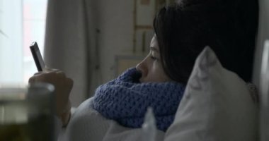 Hasta Kadın Smartphone Kullanıyor İnternet Tarayıcı Dokunmatik Ekran ve öksürük Yatak Odası 4K 'da Pencere önünde uzanırken