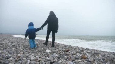Anne ve Oğlu Fırtına Denizi 'ni izlerken Taşlı Deniz kıyısında yürüyor. Dalgalar Pebble Sahili 'ne çarpıyor. Atlantik Okyanusu. Gri Bulutlu Gökyüzü. İngiliz kanalı. Fransa Avrupa. 2x Yavaş çekim 60 fps 4K