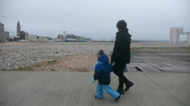 Anne ve Oğlu Deniz kenarında Yürüyen Çakıl Sahili. Coastline 'daki şehre doğru. Gri Bulutlu Gökyüzü. Le Havre Normandiya Fransa Avrupa. 2x Yavaş çekim 60 fps 4K