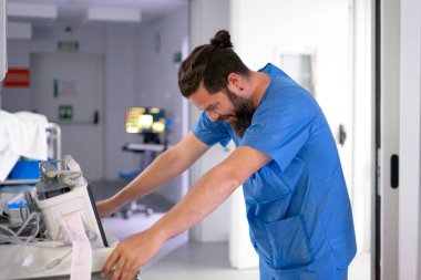 Aşırı çalışan erkek hemşire hastane koridorunda kendini yorgun ve stresli hissediyor. EKG makinesine yaslanıyor.