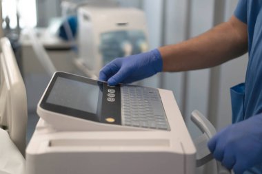 Doktorun elinde mavi eldiven, hastane odasında tıbbi cihaz kontrol paneli var.