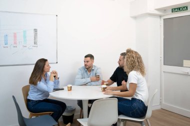 Modern bir ofis ortamında masa etrafında beyaz tahtayla işbirliği yapan dört iş arkadaşı.