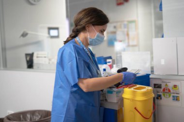 Mavi önlük, maske ve eldiven giyen sağlık çalışanı modern hastane ortamında eşyalar tarıyor.