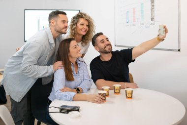 Neşeli çeşitli meslektaşlar modern ofiste kahve molası sırasında selfie çekiyorlar.