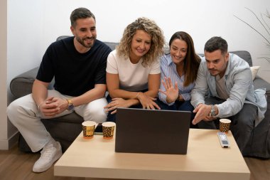 Dört arkadaş kanepede oturup dizüstü bilgisayarda video sohbeti yapıyor ve uzaktan selam veriyor.