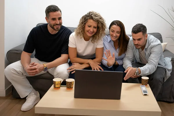 Dört arkadaş kanepede oturup dizüstü bilgisayarda video sohbeti yapıyor ve uzaktan selam veriyor.