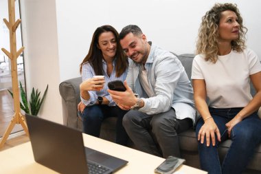 İş yerindeki bir ofis molasında koltukta gülümseyen ve akıllı telefon kullanan iş arkadaşları.