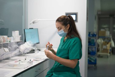 Ameliyat önlüğü ve maskeli genç bayan hemşire hastane ortamında şırıngayla ilaç hazırlıyor.