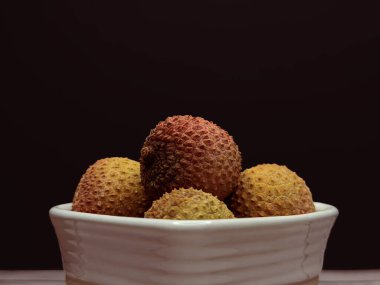 Lychee meyvesini siyah arkaplanlı beyaz kâseye koy. Tropikal desenli kabuk ve ılık tonlarda temiz, egzotik bir yiyecek, natürmort yaşam konsepti oluşturur..