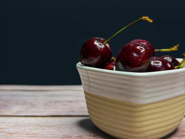 Ahşap bir masada dokulu beyaz bir seramik kasede parlak, olgun kırmızı kirazlar. Huysuz ışık ve karanlık arka plan kırsal bir yiyecek oluşturur..