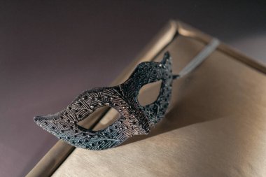 Altın çerçeveli siyah bir maske bir kitabın üzerinde duruyor. Maske süslenmiş ve altın bir süslemesi var, ona zarif ve gizemli bir görünüm veriyor. Sahne bir entrika ve cazibe hissi uyandırıyor.