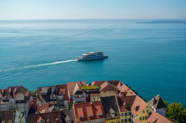 Almanya 'da Bodensee Gölü' nde renkli evlerin yakınında büyük bir gemi hareket ediyor. Sahne sakin ve huzurlu, su ve gökyüzü güzel bir ahenkle harmanlanmış.