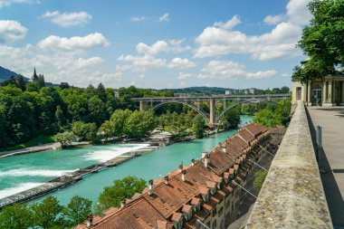 İsviçre, Bern 'deki Aare Nehri' ne bakan tepenin manzarası. Arka planda bir demiryolu köprüsü görünür. Nehir kıyısında birçok çıkıntı var. Kırmızı kiremitli çatıları olan birkaç ev.