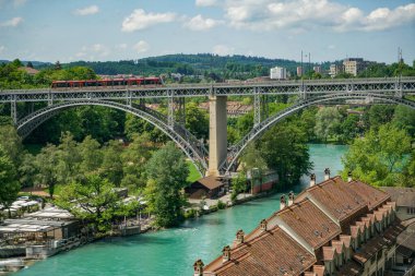 İsviçre, Bern 'deki Aare Nehri' ne bakan tepenin manzarası. Arka planda bir demiryolu köprüsü görünür. Nehir kıyısında birçok çıkıntı var. Kırmızı kiremitli çatıları olan birkaç ev.