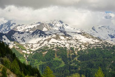Karla kaplı bir dağ manzarası. Aşağıda yeşil ağaçlar görülebilir. Gökyüzünde epeyce bulut var..
