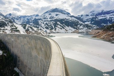 Finhaut İsviçre Alplerinde büyük bir beton baraj Tyrolienne Emossionnelle. Suda buz var. Arka planda karla kaplı dağlar görünüyor.