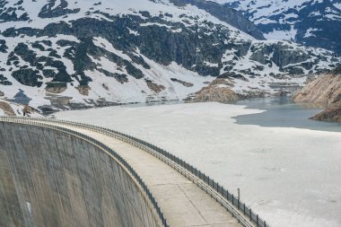 Finhaut İsviçre Alplerinde büyük bir beton baraj Tyrolienne Emossionnelle. Suda buz var. Arka planda karla kaplı dağlar görünüyor.
