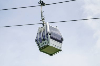 Küçük beyaz ve gümüş bir kutu bir kablodan askıya alınır. Kutu açık ve penceresi var. Gökyüzü açık ve güneş parlıyor.