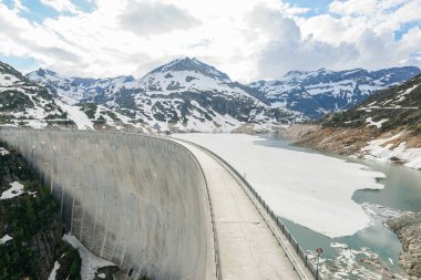 Finhaut İsviçre Alplerinde büyük bir beton baraj Tyrolienne Emossionnelle. Suda buz var. Arka planda karla kaplı dağlar görünüyor.