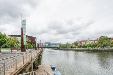 İspanya 'nın Bilbao kentindeki Nervion Nehri manzarası. Arka planda dağlar ve köprünün bir kısmı görülebilir. Tasarımcı direkleri sol tarafta görünür. Gökyüzü bulutlu ve bulutlu.