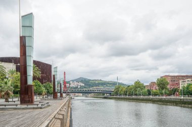 İspanya 'nın Bilbao kentindeki Nervion Nehri manzarası. Arka planda dağlar ve köprünün bir kısmı görülebilir. Tasarımcı direkleri sol tarafta görünür. Gökyüzü bulutlu ve bulutlu.