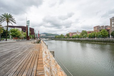 İspanya 'nın Bilbao kentindeki Nervion Nehri manzarası. Arka planda dağlar ve köprünün bir kısmı görülebilir. Tasarımcı direkleri sol tarafta görünür. Gökyüzü bulutlu ve bulutlu.