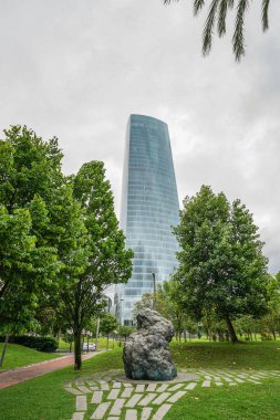 İspanya 'nın Bilbao şehrinde bulutlu bir gökyüzüne karşı yüksek bir gökdelen. Ön planda, tanımlanamayan bir şekle sahip bir heykel görünür..