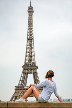 Kısa saçlı, şort ve beyaz ayakkabılı mavi bluzlu bir kız Paris, Fransa 'da arka planda Eyfel Kulesi olan bir kayanın üzerinde oturuyor. Akşamları hava bulutlu..