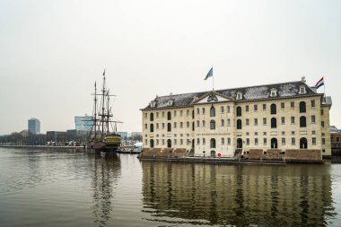 Amsterdam Nakliye Müzesi binasının yanında eski bir ahşap yelkenli gemi görülüyor. Fotoğraf bulutlu ve kasvetli bir kış gününde çekildi..