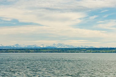 Almanya, Bodensee Gölü 'nden İsviçre Alpleri' nin panoramik görüntüsü. Su sakin, gökyüzü mavi, hava güneşli, gökyüzü bulutlarla kaplı..