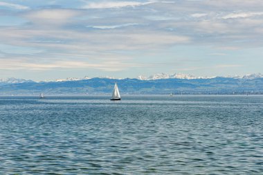 Arka planda açık mavi gökyüzü olan bir yelkenli göle açılıyor. Gökyüzü bulutlu, ama güneş hala bulutların arasında parlıyor. Suda görünen tek tekne yelkenli.