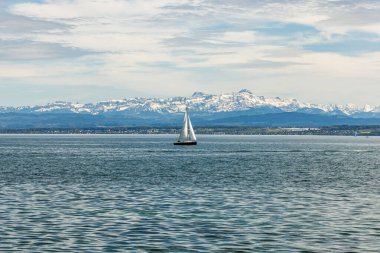 Arka planda açık mavi gökyüzü olan bir yelkenli göle açılıyor. Gökyüzü bulutlu, ama güneş hala bulutların arasında parlıyor. Suda görünen tek tekne yelkenli.