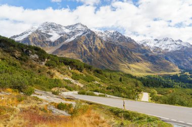 İsviçre 'deki San Bernardino Geçidi' nde asfalt bir yol. Dağlar kısmen karla kaplı. Dağlar da güzel, renkli bir sonbahar battaniyesiyle kaplıdır. Gökyüzü mavi ve bulutludur..