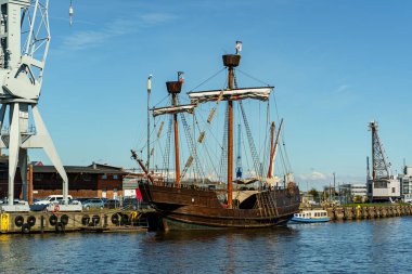 Eski bir ahşap yelkenli gemisi Almanya 'nın Lubeck kentindeki rıhtımda duruyor. Gri bir iskele vinci ve diğer gemileri görüyoruz..