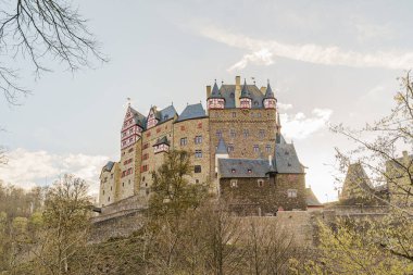 Eltz Şatosu ormandan yaklaşırken aşağıdan çekiliyor. Gökyüzü mavi ve bulutlu.